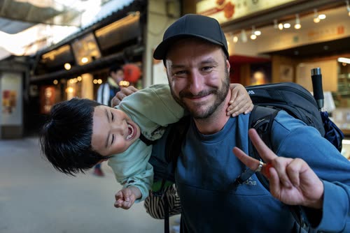 Jordy Scott carrying a laughing child on his shoulders in a covered market street
