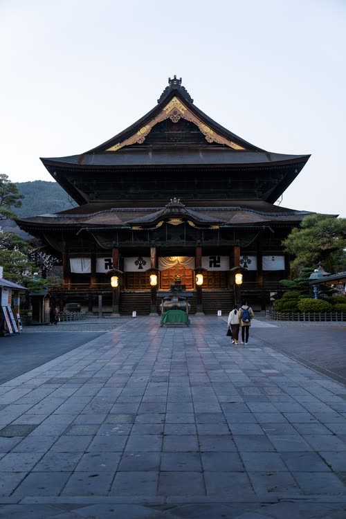 The great wooden main hall of Zenko-ji at dawn, two visitors crossing the stone forecourt