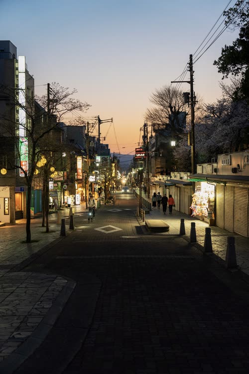 A quiet sloping street of shopfronts glowing at dusk