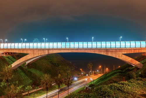 A lit road bridge arching over a green ravine in Lima at dusk