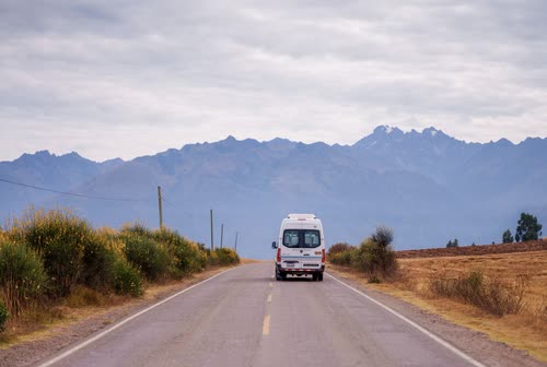 A minibus on a straight highway running toward snow-capped Andean peaks