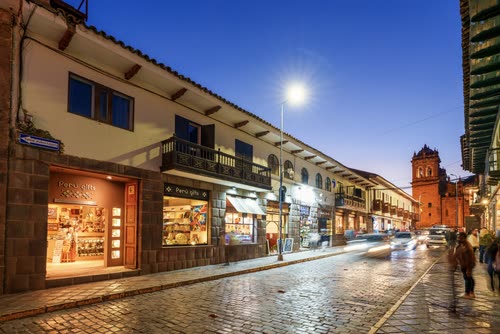 Lit shopfronts along a cobbled Cusco street at blue hour