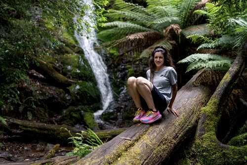 A woman sitting on a mossy log beside a fern-lined waterfall, Tasmania